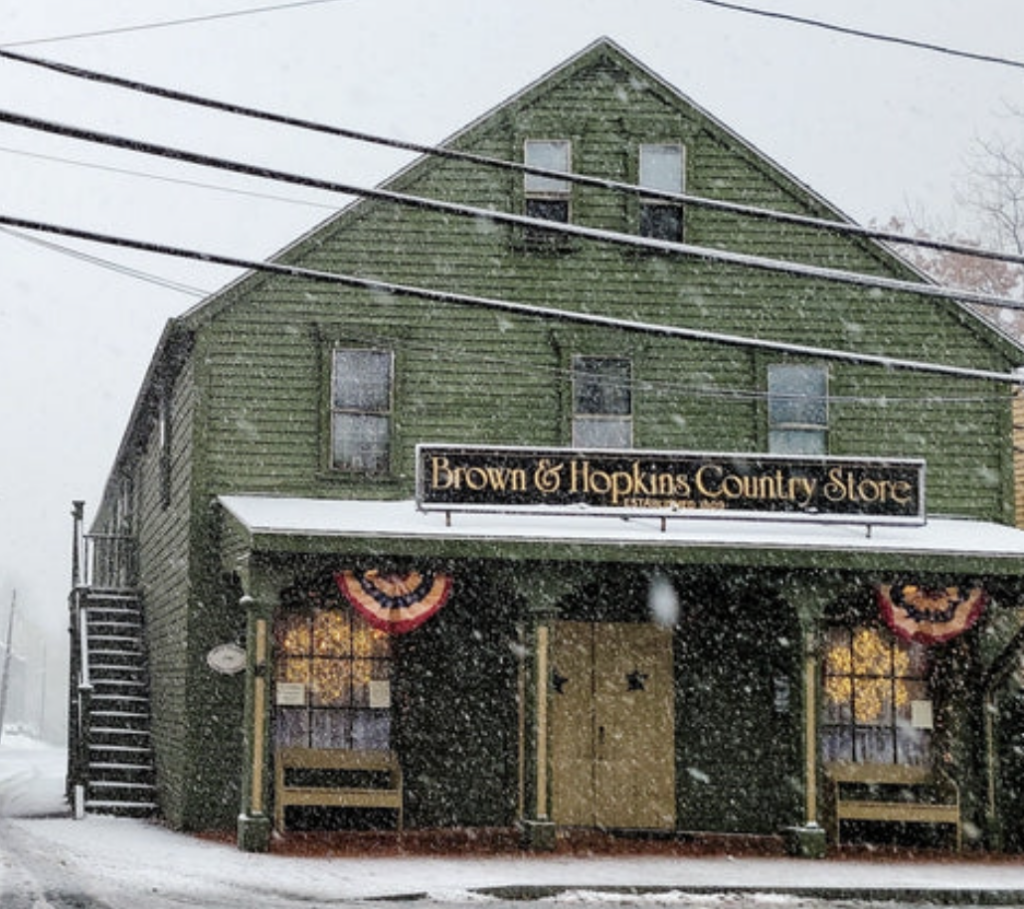 A green, snow-covered building labeled "Brown & Hopkins Country Store" with American bunting on either side of the entrance. Snow is falling, and there's an exterior staircase on the side of the building.