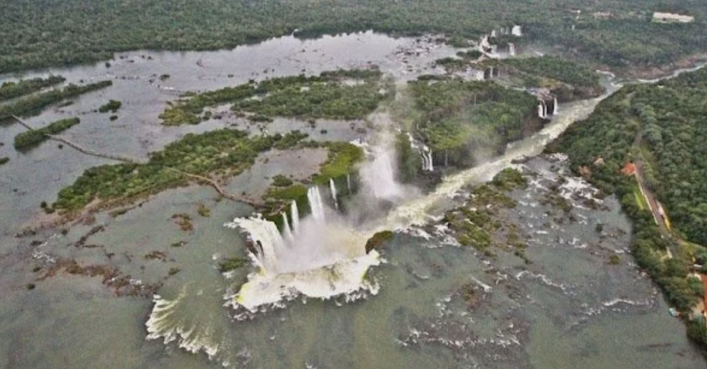 Aerial view of Iguazu Falls, showcasing a series of waterfalls cascading over lush, forested cliffs. The wide, flowing river and green vegetation surround the falls, creating a dramatic and expansive landscape.