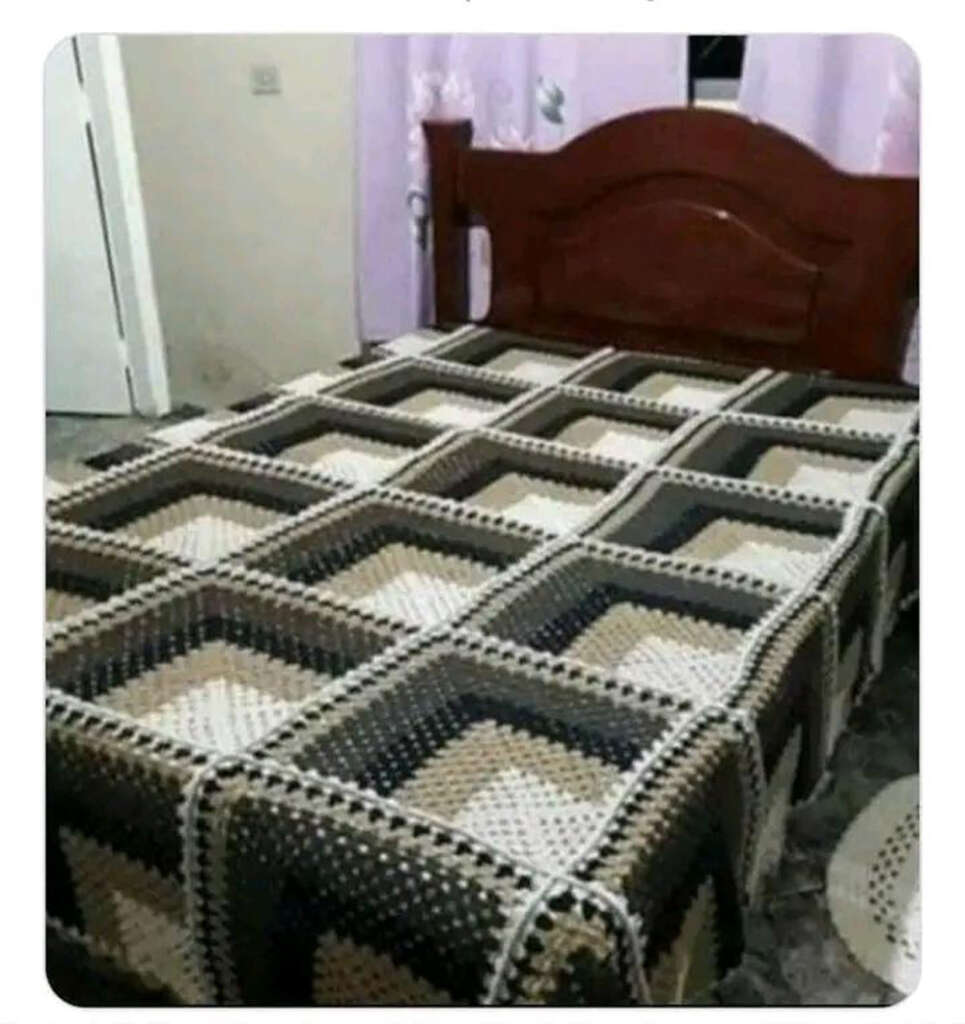 A wooden bed with a crochet blanket featuring a geometric square pattern in shades of brown, beige, and black. The room has light purple curtains and a marble-patterned floor.