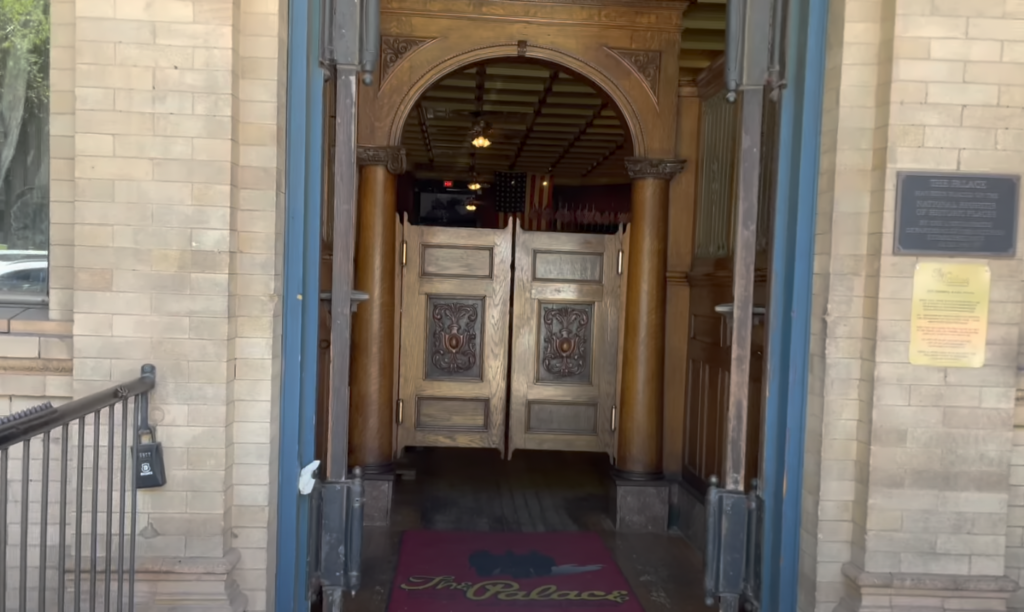 Entrance of a historic building with wooden swinging saloon doors. The doors are flanked by ornate columns and lit by warm overhead lights. A burgundy mat on the ground reads "The Palace." Plaques with text are mounted on the brick walls.