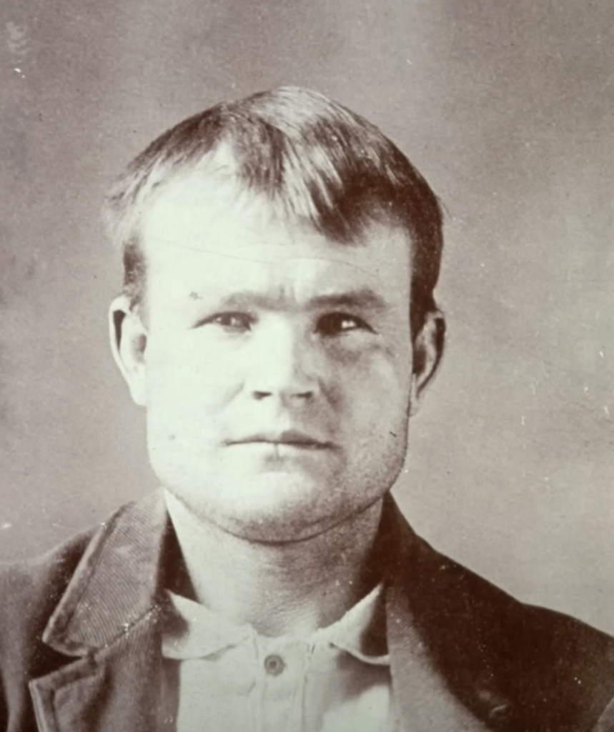 A sepia-toned vintage photograph of a man with short hair. He wears a jacket over a button-up shirt and looks directly at the camera. The background is plain, emphasizing his expression and attire.