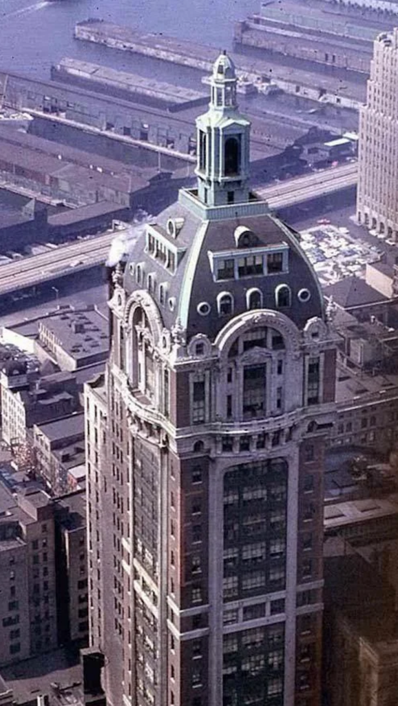 Aerial view of a historic skyscraper with a green dome and clock faces on its upper section. Surrounding buildings and a river with piers can be seen in the background, enhancing the urban setting.