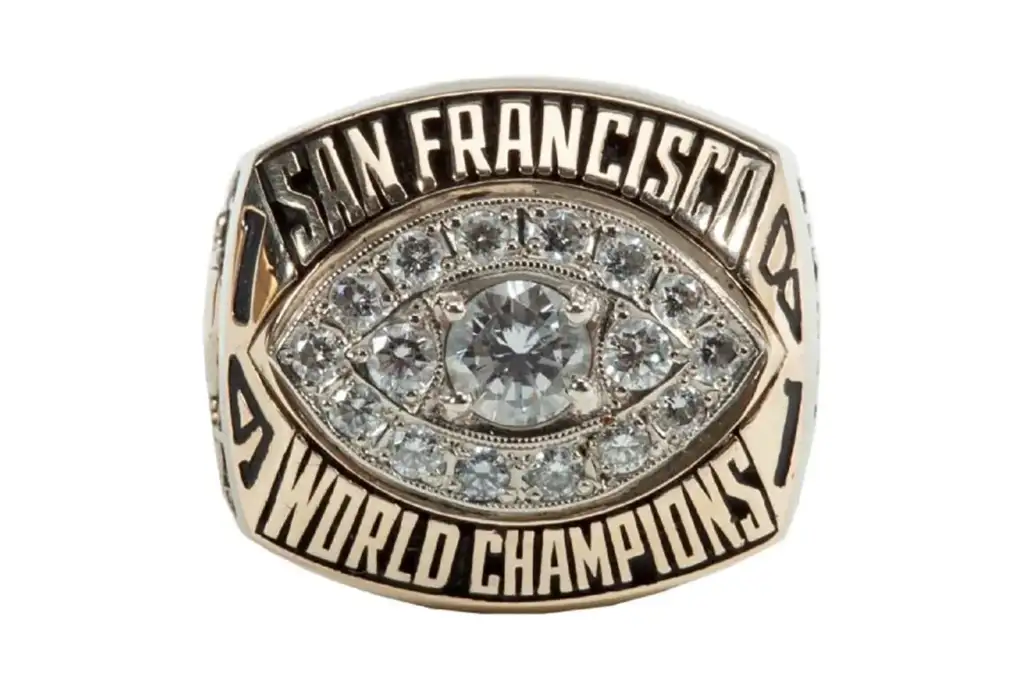 A gold ring featuring a large central diamond surrounded by smaller diamonds in a football shape. Engraved text reads "San Francisco" at the top and "World Champions" at the bottom.