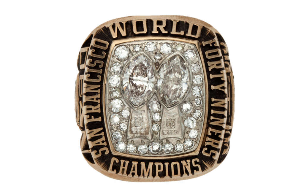 A gold San Francisco 49ers championship ring with "World Champions" inscribed. It features two large diamond-shaped stones surrounded by smaller diamonds and intricate details, symbolizing their football victories.