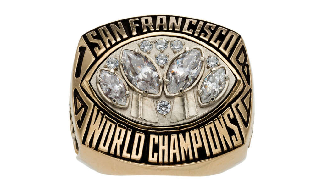 A gold championship ring with bold black lettering that reads "San Francisco World Champions." The front is adorned with multiple large and small gemstones.