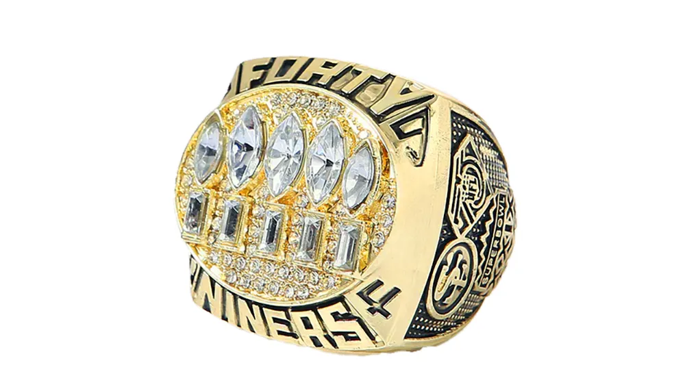 A gold championship ring featuring large, diamond-like stones on top, surrounded by intricate patterns. The words "49ERS" and "WINNERS" are engraved on the band.