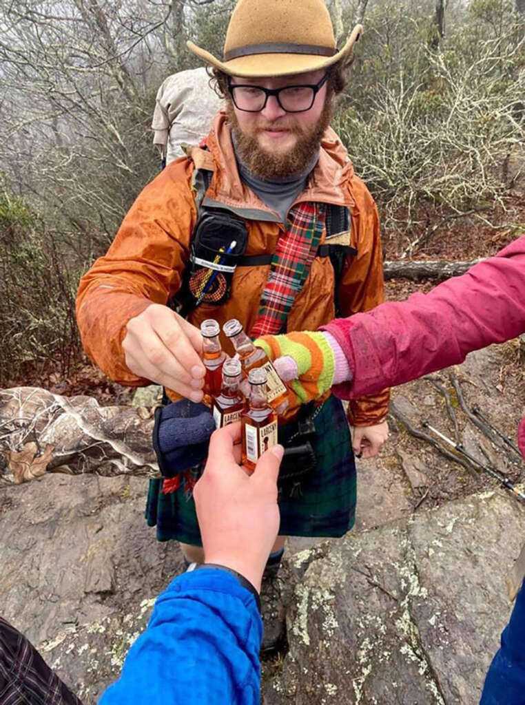 People wearing outdoor clothing and colorful sleeves are toasting with small bottles on a rocky trail. One person, wearing a kilt and hat, holds multiple bottles, while others extend their hands to clink them together. Sparse trees are in the background.