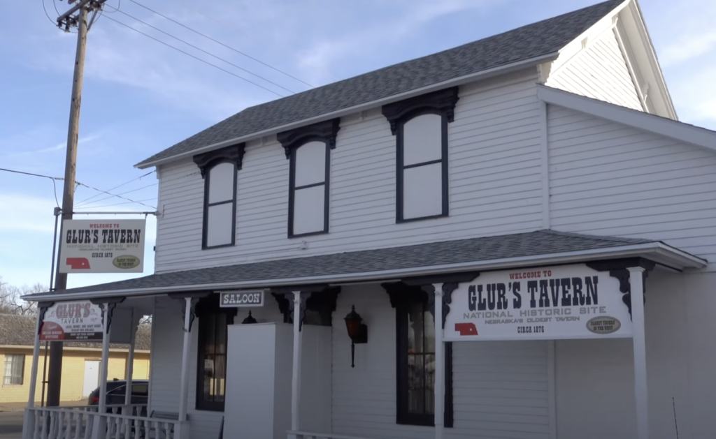 A white, two-story building with black trim, featuring signs that read "Glur's Tavern" and "National Historic Site." The structure has a small front porch and is under a clear blue sky.