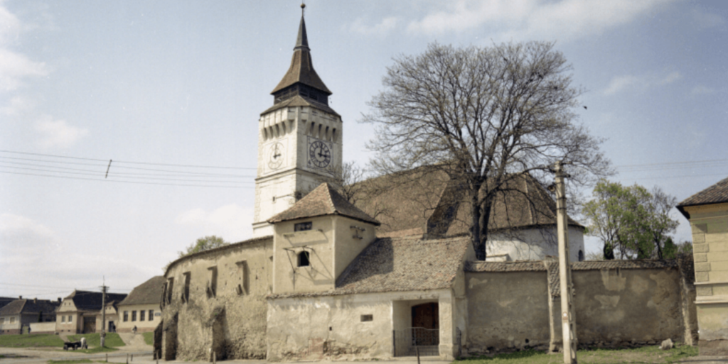 A historical church with a tall clock tower rises behind ancient stone walls. The building is flanked by leafless trees, and a few surrounding houses are visible under a partly cloudy sky.