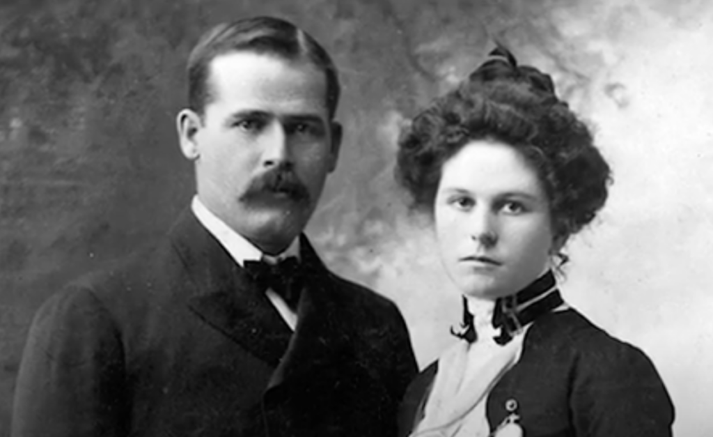 A vintage black and white portrait of a man with a mustache wearing a suit and bow tie, standing next to a woman with her hair styled up, wearing a high-collared dress with buttons. Both are looking directly at the camera.