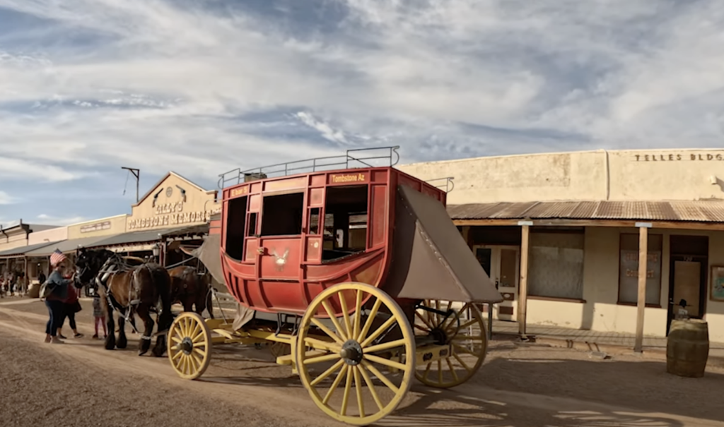 A red stagecoach with yellow wheels is parked on a dusty street in a Western-style town. Two horses are hitched to the stagecoach. People walk nearby, and the background shows rustic wooden buildings under a partly cloudy sky.