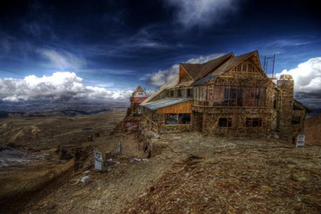 A rustic stone building sits atop a barren, rocky mountain under a dramatic, cloudy sky. The landscape stretches into the distance, with patches of snow visible on the ground. The atmosphere is moody and remote.