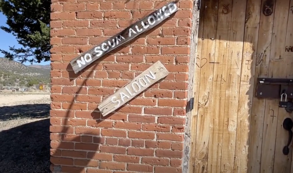 Brick wall with wooden signs reading "NO SCUM ALLOWED" and "SALOON." A wooden door is partially visible to the right. The area is outdoors, with trees and a clear sky in the background.