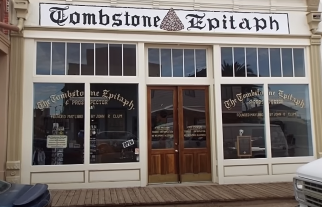 Front view of a historic building with a sign reading "Tombstone Epitaph." The facade includes large wooden doors and windows displaying newspaper-related content. Text on the windows highlights "The Tombstone Epitaph" as a historic publication.