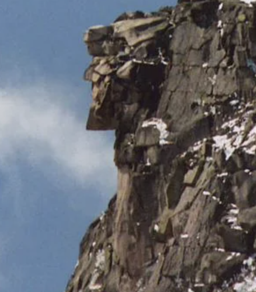 Rock formation resembling a profile face, with distinct nose and chin features, on a cliff. The rock surface is jagged with some snow patches. A clear blue sky is in the background.