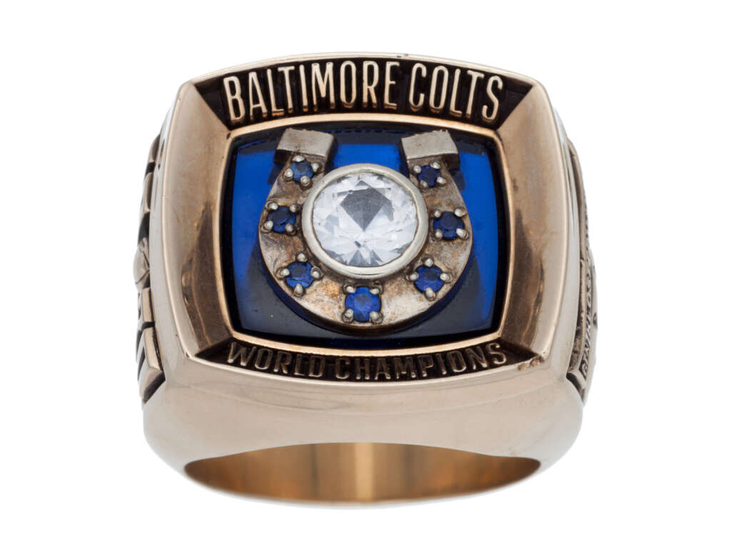 Gold Baltimore Colts championship ring with a large diamond center, surrounded by a horseshoe design with blue stones. "World Champions" is engraved beneath the horseshoe on a blue background.