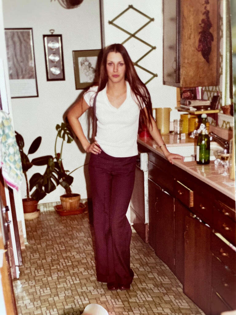 A woman with long hair wearing a white shirt and dark pants stands in a retro-style kitchen. She rests one hand on the counter amidst various kitchen items. Potted plants are visible in the background on patterned flooring.