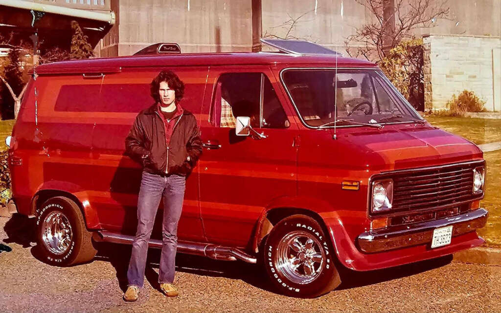 A person stands in front of a vintage red van with custom stripes. They are wearing a black jacket and jeans, with hands in pockets. The background includes a building and some trees, suggesting a suburban setting.