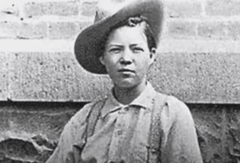 A person wearing a cowboy hat and a button-up shirt is looking at the camera. They are standing in front of a textured stone wall. The image is in black and white, giving it a vintage appearance.