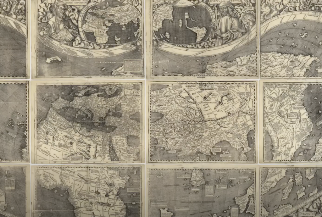 Antique black and white world map with detailed sections, each square depicting a different region. Intricate borders with artistic figures and globe illustrations are visible on the top panels.