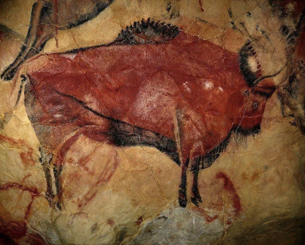 Ancient cave painting depicting a large, red bison with black outlines on a textured rock surface. The artwork features detailed lines and shading, showcasing the bison's horns and body, against a background of earthy tones.