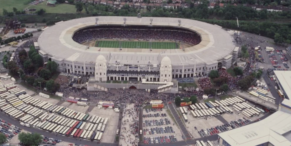 Aerial view of a large, crowded stadium with a green field in the center. The surrounding area is filled with parked vehicles and people milling around. The stadium has distinctive white towers and an open roof design. Trees and buildings are visible nearby.