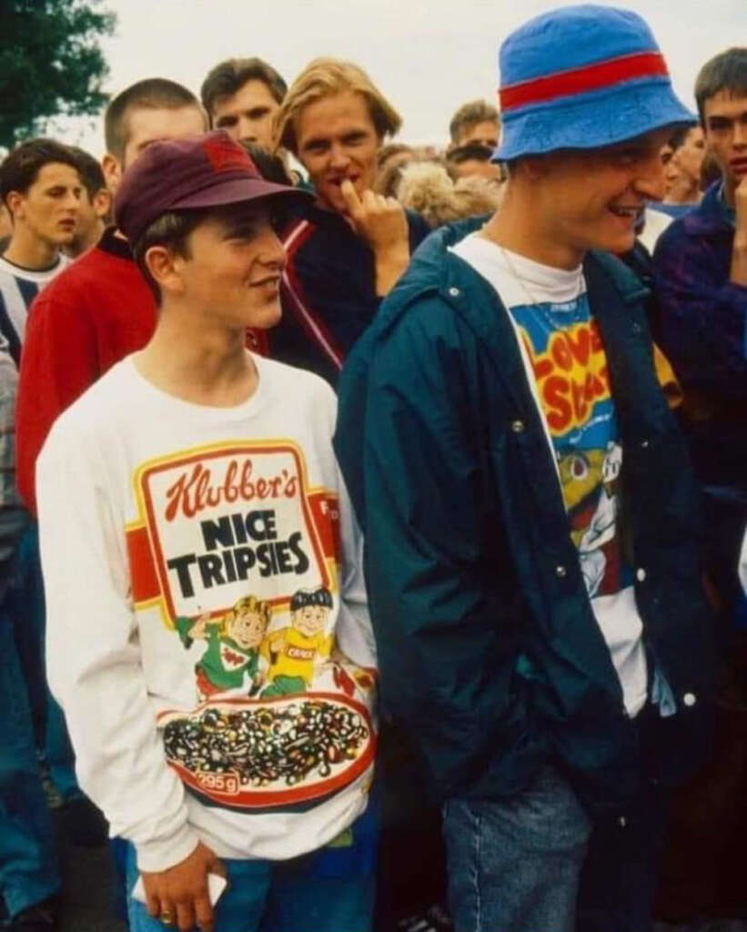 Two people stand in a crowd, smiling. One wears a maroon cap and white sweater with cartoon graphics. The other wears a blue and red hat and a dark jacket over a colorful t-shirt. More people are visible in the background.