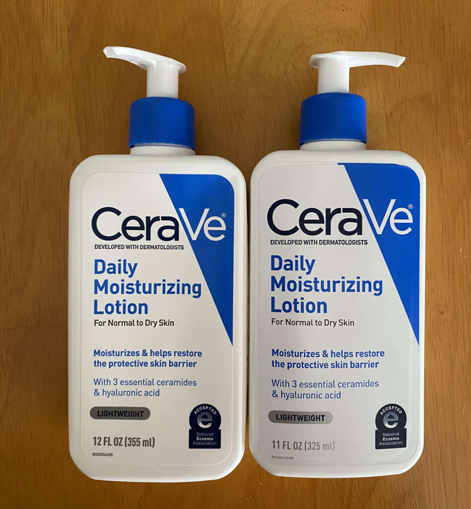 Two bottles of CeraVe Daily Moisturizing Lotion are placed on a wooden surface. Each bottle features a pump and contains 12 fl oz and 11 fl oz, respectively. The label highlights ingredients like ceramides and hyaluronic acid.