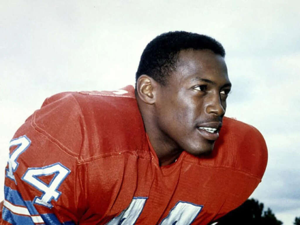 A football player wearing a red jersey with the number 44 crouches while looking to the right. The jersey has blue and white stripes on the sleeves. The sky is cloudy in the background.