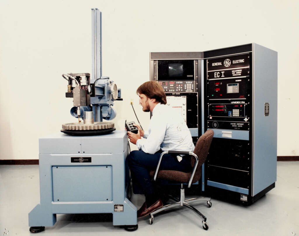 A person seated on a chair operates a large industrial machine with controls and a computer system labeled "General Electric EC-I." The equipment is in a clean room with a light-colored floor and walls.