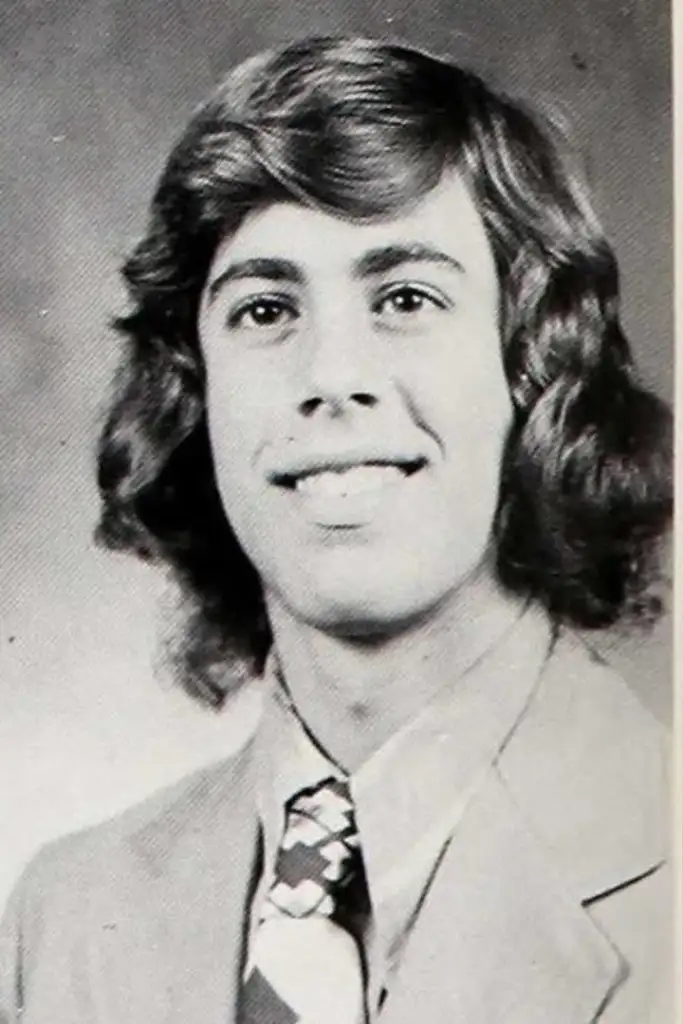 A vintage black and white yearbook photo of a young person with medium-length, wavy hair. They are wearing a suit jacket and a patterned tie, smiling at the camera.