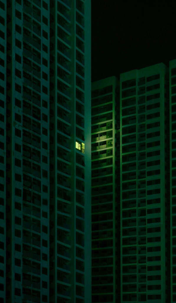 Tall, dark green buildings at night with one lit window illuminating part of the facade. The scene is dim, emphasizing the contrast between the illuminated window and the surrounding darkness.