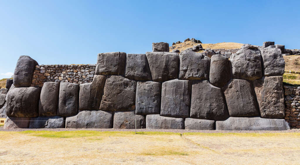 Massive stone walls with large, closely-fitted blocks set against a clear blue sky. The ancient structure shows meticulous stonework, possibly Incan. Dry grass covers the foreground, and some rocky terrain is visible in the background.