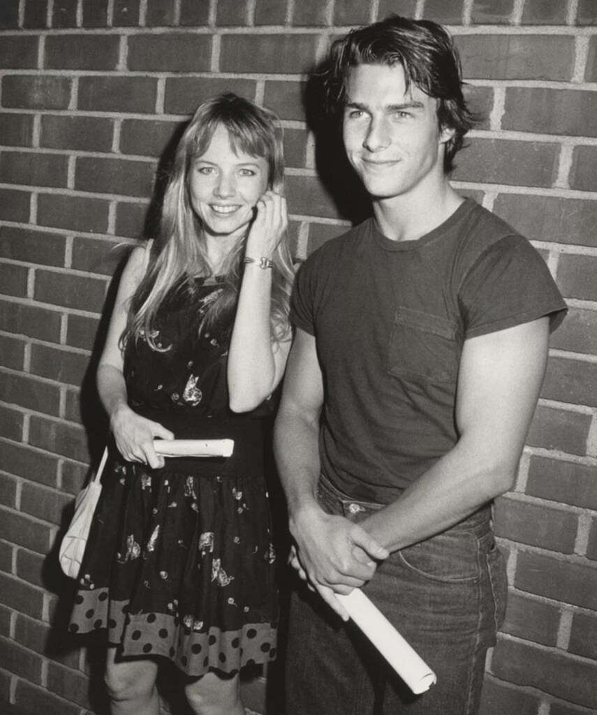 A young woman in a floral dress and a young man in a T-shirt and jeans stand together, smiling, against a brick wall. They both hold rolled-up papers. The photo is in black and white.