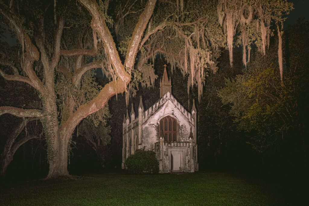 A small, illuminated chapel stands in a dark, forested area, with large trees draped in Spanish moss surrounding it. The Gothic architecture and night setting create an eerie atmosphere.