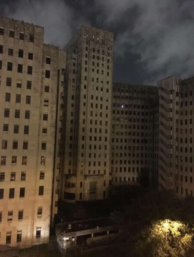 A tall, abandoned building with a weathered exterior against a cloudy night sky. The structure features numerous windows, and the area is dimly lit by a yellow streetlight near the bottom edge.