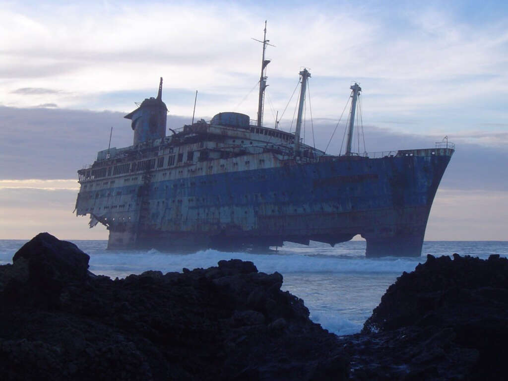 An old, rusty shipwreck rests partially submerged in the ocean near rocky shorelines under a cloudy sky. The silhouette of the ship is prominent against the backdrop of a dim, overcast horizon.