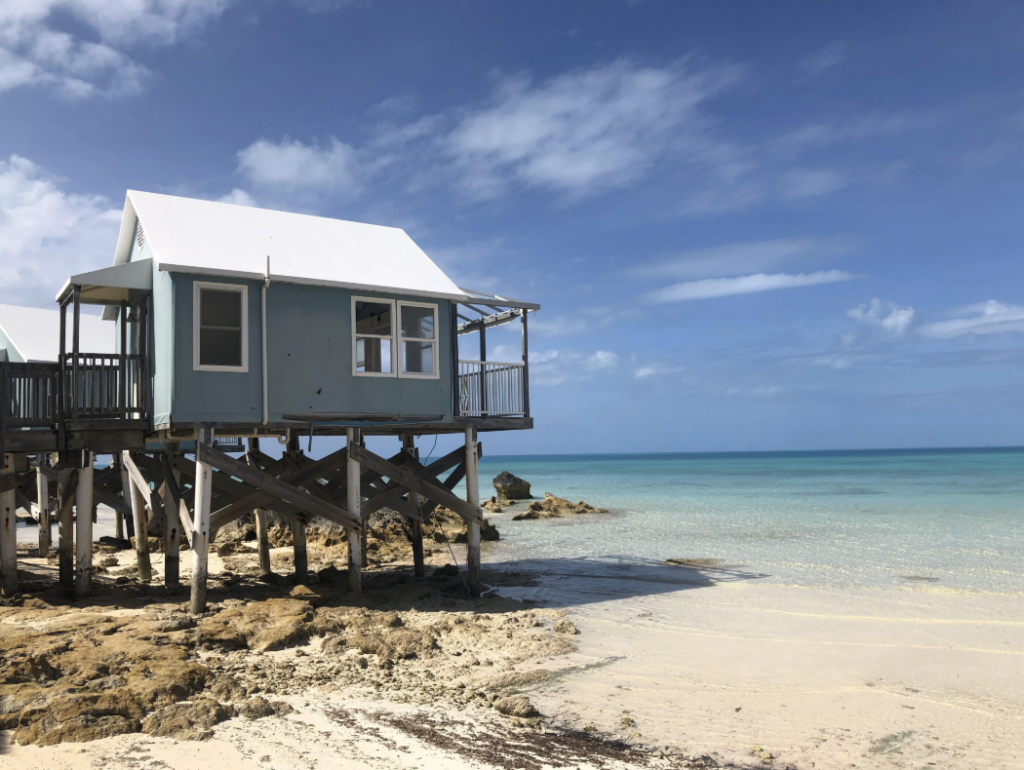 A small blue house on stilts stands on a rocky beach with clear, shallow water. The sky is bright blue with a few clouds, and the ocean extends to the horizon. The scene is tranquil and sunny.