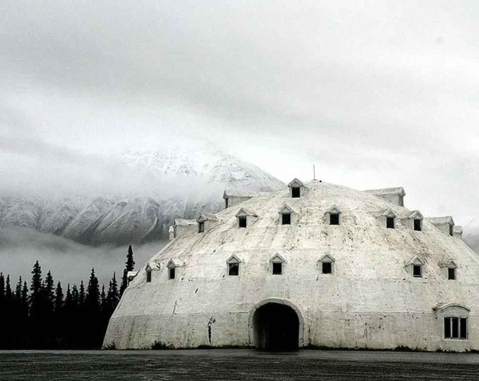 A large, dome-shaped igloo building with small triangular windows stands against a backdrop of snow-covered mountains and evergreen trees. The sky is overcast and the area appears deserted.