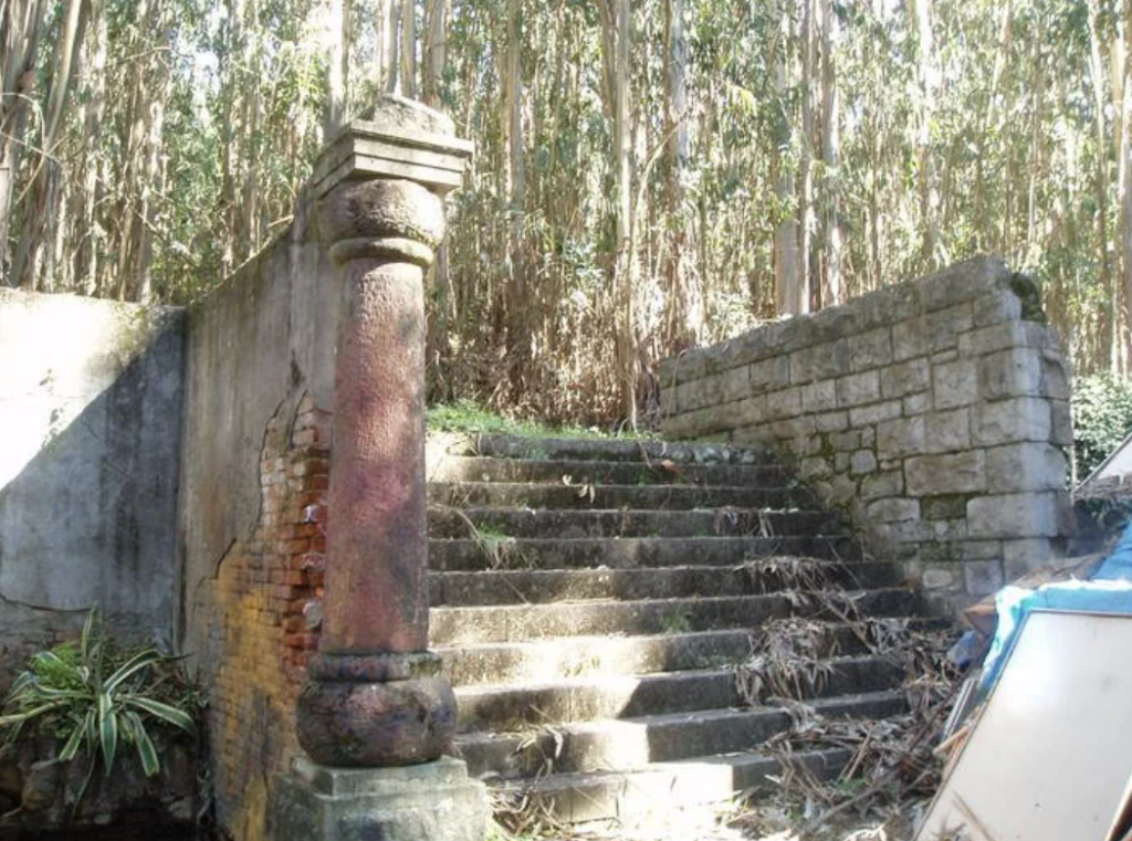 Stone steps leading up, flanked by a tall, weathered column and a brick wall on the left, with a stone wall on the right. Surrounded by dense trees and vegetation, it appears as an old, abandoned structure in a forest setting.