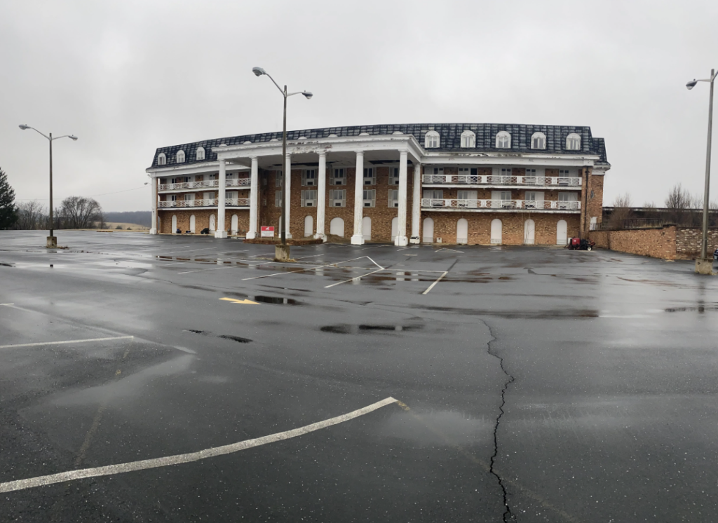 A large, mostly empty parking lot in the foreground leads to a two-story brick building with a mansard roof and tall white columns. The sky is overcast, and the ground appears wet from rain.