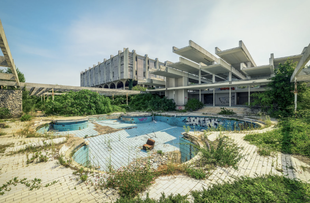 Abandoned, overgrown swimming pool with graffiti on the dry basin. The surrounding area is covered with weeds and the backdrop features a large, deteriorating concrete building with multiple levels and exposed beams under a cloudy sky.