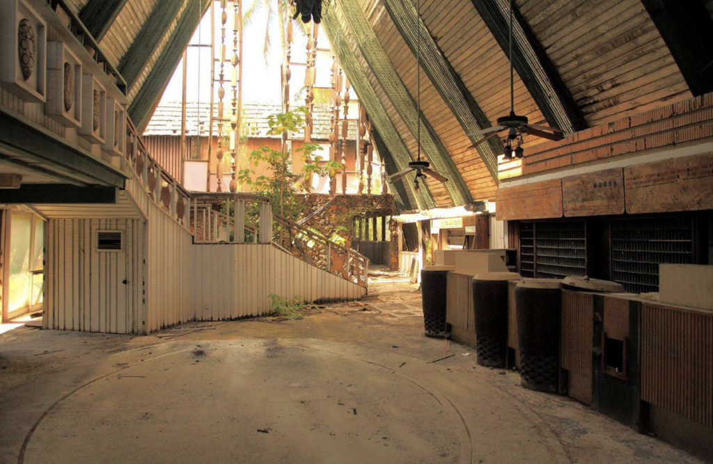 Abandoned building interior with high, sloped wooden ceilings and overgrown plants. Dusty counters and deserted reception area are visible, along with a staircase on the left and sunlight streaming through broken windows.