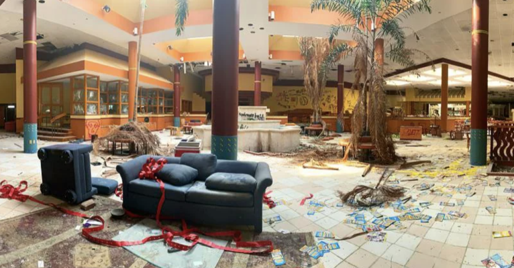 Abandoned shopping mall interior with a blue couch and suitcase surrounded by debris, fallen ceiling tiles, plants, and scattered papers. A red ribbon is draped over the couch, and palm trees grow inside the derelict space.