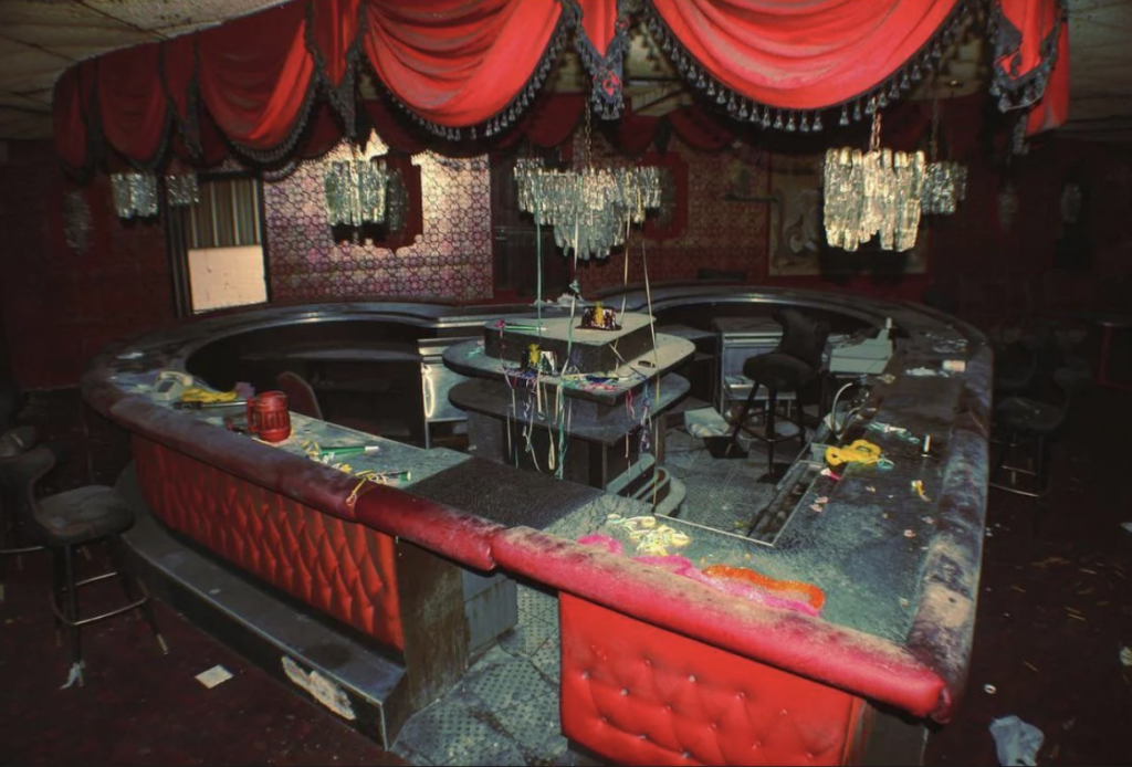 A dimly lit, abandoned bar with a curved counter, red quilted siding, and draped red curtains. Dust covers the surfaces, chandeliers hang above, and scattered items lie around, suggesting neglect and disuse.