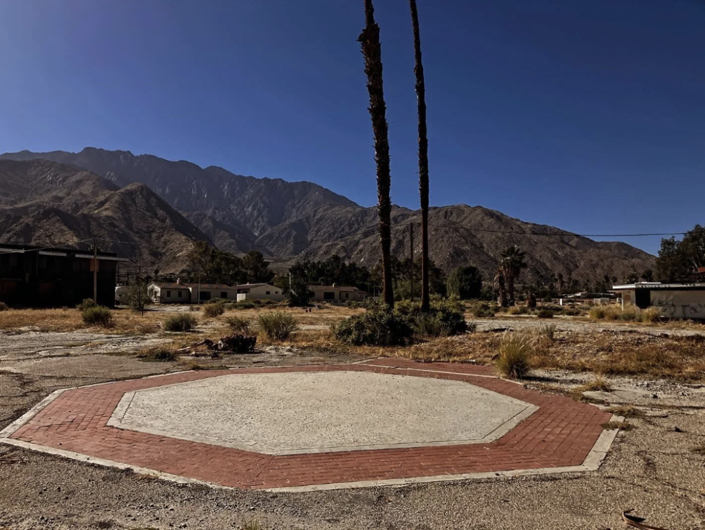 Abandoned hexagonal fountain area with two tall palm trees against a backdrop of dry mountains and clear blue sky. The ground is barren with patches of dry grass and scattered buildings.