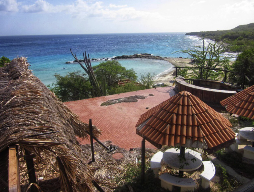 View of a coastal area with a clear blue sea and curved shoreline. There are thatched roofs and red-tiled structures in the foreground surrounded by greenery. A sandy beach is visible along the water's edge under a partly cloudy sky.