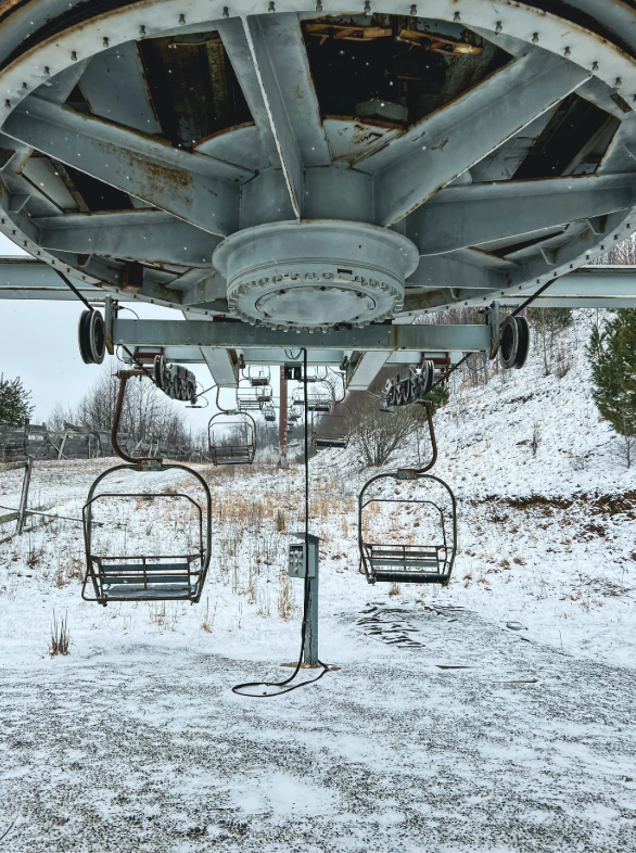 A snowy landscape with an old, abandoned ski lift system. Two rusted, empty chairlift seats hang from the mechanism. The ground is lightly covered with snow, and trees are visible on a hill in the background. The scene is overcast and desolate.