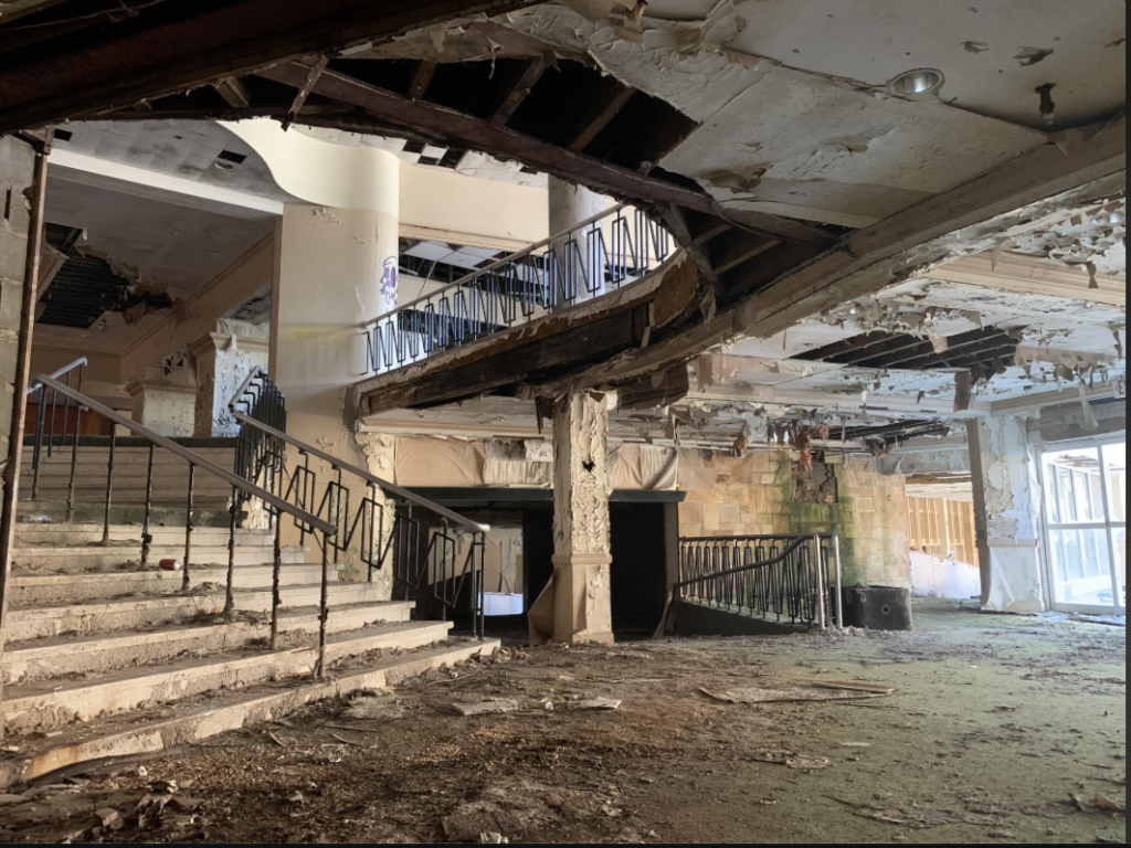 An abandoned, dilapidated building with a partially collapsed ceiling. Stairs lead up, flanked by rusting railings. Debris and dirt cover the floor, and exposed beams are visible. The atmosphere is one of decay and neglect.