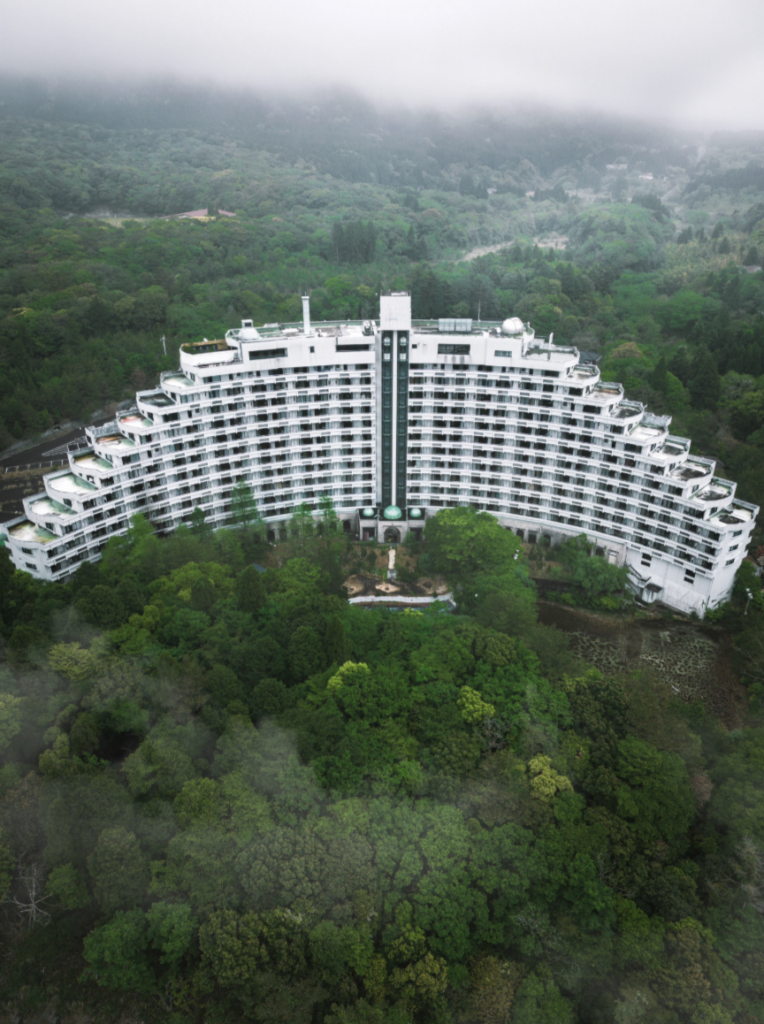 Aerial view of a large, white, crescent-shaped hotel surrounded by dense, green forest. The hotel has multiple balconies and a central black vertical stripe. Mist partially obscures the forested hills in the background.